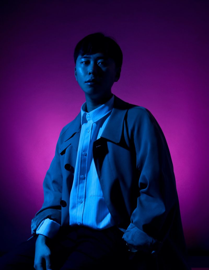 A man standing in a white shirt and overcoat in front of a purple light on a black wall.